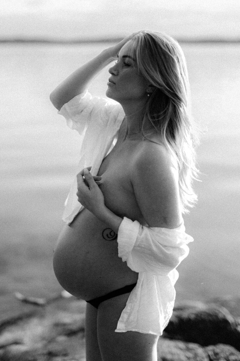En gravid kvinna ikädd vit skjorta ståendes vid vattnet med ena handen i håret under hudnära gravidfotografering i Stockholm