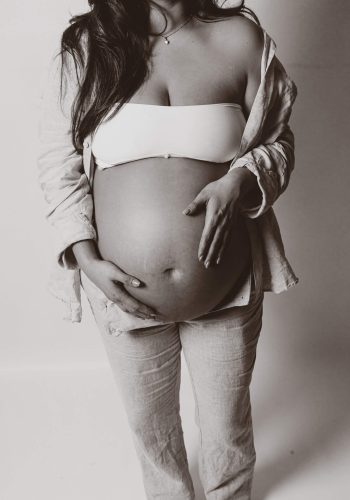 En gravid kvinna ståendes med båda hennes händer på sin gravida mage under en gravidfotografering i studio Stockholm