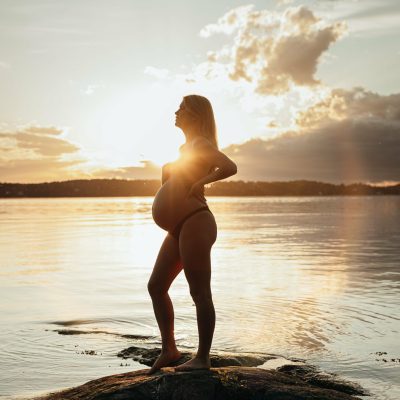 Gravid kvinna står på en sten i vattnet med solen i bakgrunden